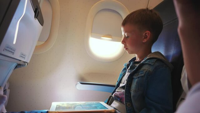 Little boy playing video games on tablet looking at screen and touching finger during flight in airplane. Child tourist traveling by plane having digital gadget for entertainment or educational app.