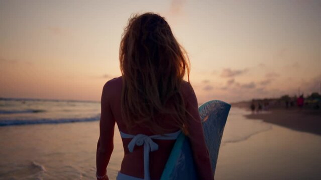 Woman surfer with surf on ocean beach. Blonde girl enjoying sunset on resort touching hair after surfing holding surfboard in hands on sandy shore. Sports outdoors activity tourism rest relax concept.