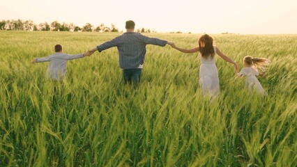 Happy family with child runs through wheat field holding hands. Slow motion. Mom, dad and children are walking together. Cheerful mother, father and little daughter play, enjoy nature outdoors, dream