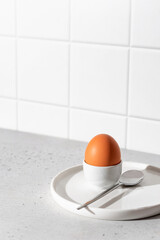 Boiled egg in an egg stand and spoon on white tile kitchen background with text space. Light...