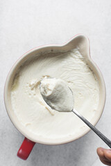 Scooping out homemade greek yogurt in a ceramic bowl with a spoon, process of making Greek yogurt, strained thick yogurt in white mixing bowl