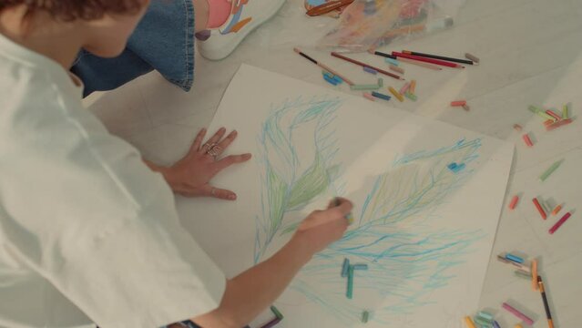 Top shot of hands of unrecognizable female artist in beanie hat and white t-shirt sitting on floor and drawing botanic picture with multicoloured pastel and pencils