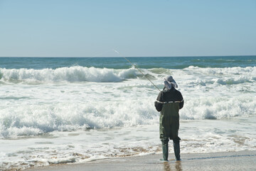 A sea angler covered in sun protection gear stands on the beach facing the rolling waves