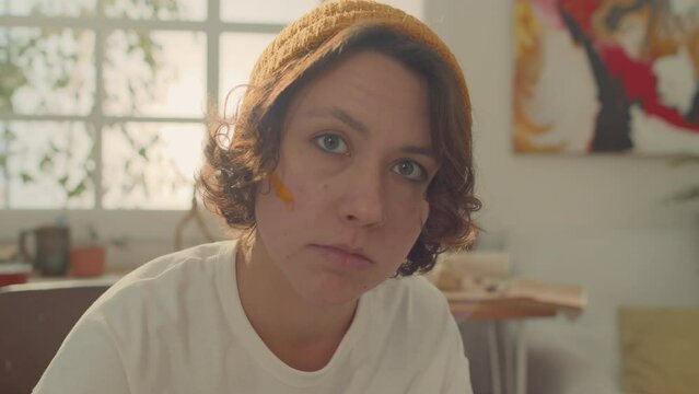 Close-up portrait shot of face of young curly biracial woman in beanie hat, white t-shirt, with nose ring sitting in front of window at home or in art workshop, turning head and looking at camera