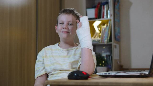 Boy at table with ironic smile shows cast on his arm and cannot use computer. Acceptance and grief due to limitations in activities of child with hand injury. Portrait of teenager at home at table.