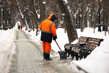 Communal services worker in uniform with a shovel clears snow on a sidewalk. Man during snow...