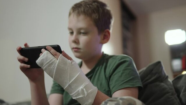 Focused child with fracture and cast on his arm watches video on smartphone and is surprised. Carefully watch online video by subscription with hand injury during recovery at home.