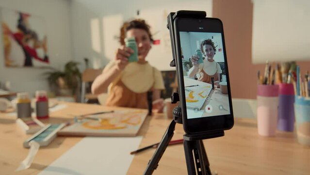 Close-up shot of smartphone screen filming Caucasian female artist sitting at table in workshop, talking to camera, demonstrating pot of paint, while recording lesson or tutorial for internet blog