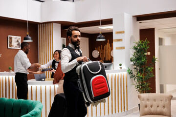 Hotel porter helping tourists with bags, carrying suitcases to accommodation in modern resort. Young bellhop working as steward providing luxury service to guests with luggage at front desk.