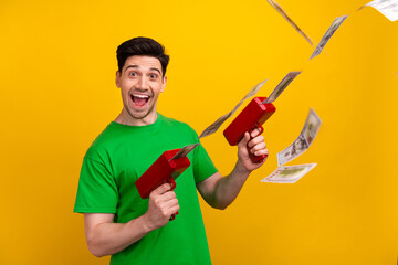 Photo of funky successful man with stylish haircut dressed green t-shirt hold guns shooting money isolated on yellow color background