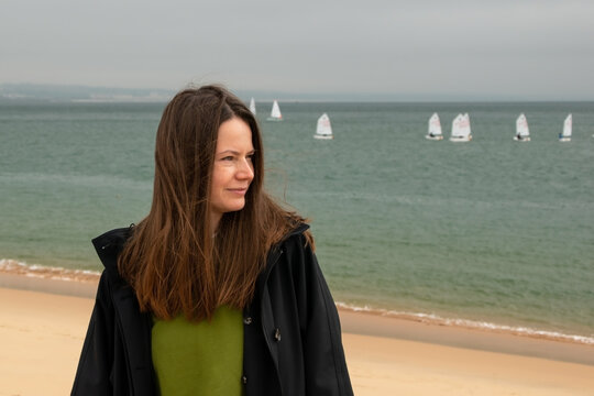 Young woman standing on the beach with sailing boats in the background.