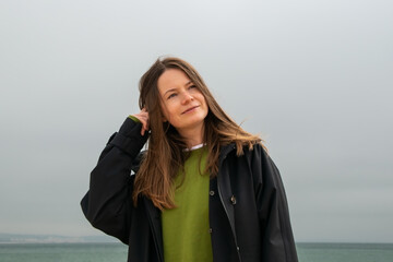 Young woman with long brown hair, wearing a black leather jacket and a green T-shirt, standing in front of the sea on a cloudy day.