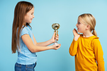Portrait of beautiful little girl giving triumph cup to her sister, standing on blue background