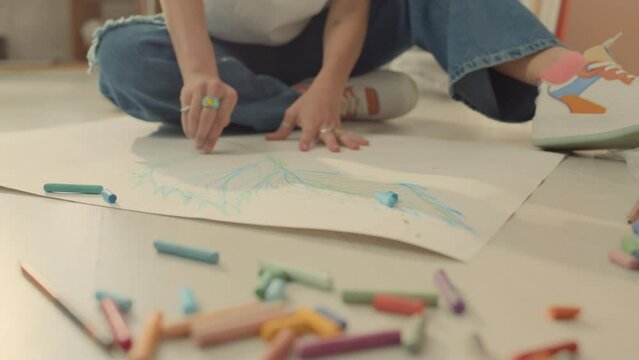 Close-up shot of hands of unrecognizable woman or girl in jeans, trainers sitting on floor in art class or at home, and drawing picture on paper with colourful chalk or crayons