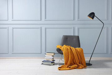 Comfortable armchair with blanket, books and lamp near grey wall indoors, space for text
