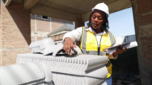African American black woman architect in hard hat holding digital tablet and blueprints examining roof tiles using house under construction, housing construction project area