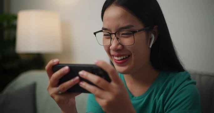 Footage of Young Asian woman wearing glasses and headphones enjoys playing online esport games on smartphone sitting on sofa in the living room at home,Gamer lifestyle concept.