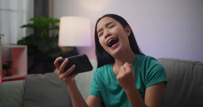 Footage of Young Asian woman enjoys playing online esport games on smartphone sitting on sofa in the living room at home,Gamer lifestyle concept.