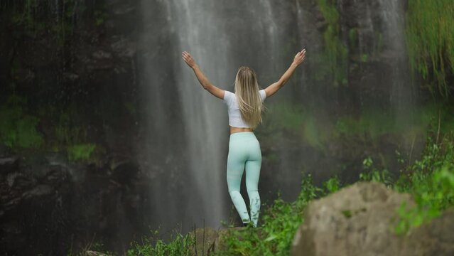 Fit woman in sportive clothing reaching base of tropical waterfall throwing hands up