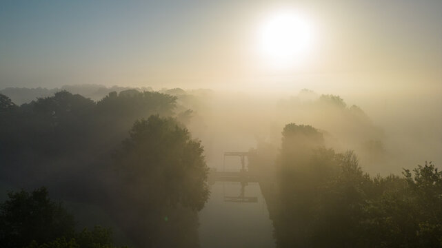 This evocative image captures the delicate balance of light and mist at dawn, with the sun rising over a tranquil river. The river itself is barely visible, as a thick blanket of fog softly engulfs