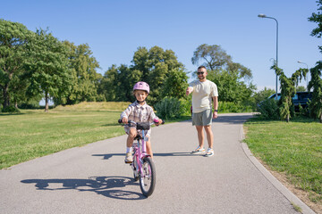 Excited father teaching girl to ride a bike, summer fun and park outdoors. Happy kid, learning and riding bicycle with help from dad, parent and safety for healthy development.