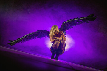 Beautiful girl an angel with the black wings posing in the smoke on dark background in the purple light. Angel of the night concept.
