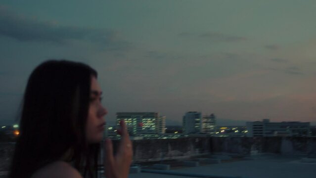 Young brunette woman's hand is raised above with cigarette smoking cigarette at rooftop with a city view