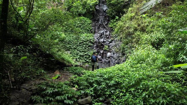 Women hiker takes in the stunning views of a rainforest waterhole and cascading waterfall along a walking track. Creative drone view