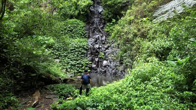 Female hiker takes in the breathtaking views of a hidden waterhole and cascading waterfall along a tourist walking track. Creative drone view