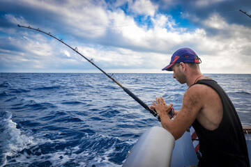 Tough man wearing baseball cap on board ship while fishing against blue sea background. - obrazy, fototapety, plakaty