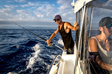 A sturdy man wearing a baseball cap aboard a boat while fishing. A girl at the helm. trolling