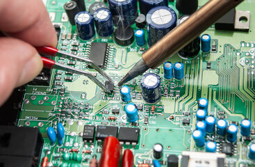Close up of a technician's hands in a workshop. Repairer is soldering circuit board of electronic device on table with tweezers. - Powered by Adobe