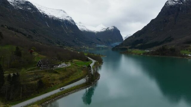 aerial view of a car moving on an asphalt road highway on the shore of Oldevatnet blue mountain lake with mountains covered with snow on background in norway