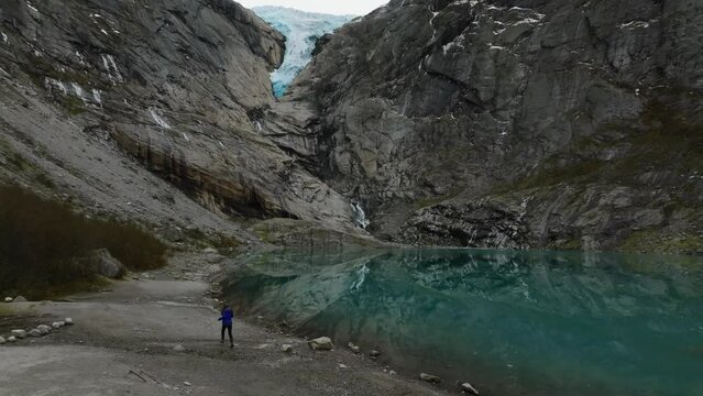 aerial view of a walking woman tourist hiker on rocky shore of briksdalsbreen glacier blue lake in norway