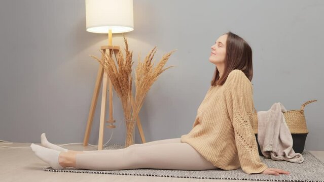 Side view of brown haired woman sitting alone on floor enjoying carefree peaceful weekend leisure time in modern stylish living room looking around dreaming