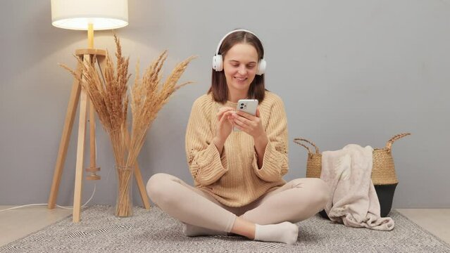 Beautiful adult Caucasian woman wearing beige jumper using smartphone and wireless headphones while sitting on floor in home interior scrolling online looking for web site for downloading music