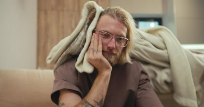 A tired blond man with a beard and glasses covered himself with a blanket and his little albino son pulls the blanket off him and wants to play with his father in a modern apartment at home