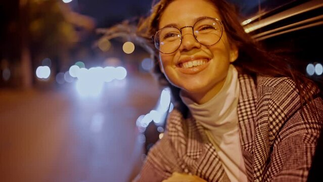 A happy and cheerful brunette girl in round glasses smiles broadly in a gray coat and looks out of the rear window of a modern car in the evening. Happy brunette girl looks out of the car window