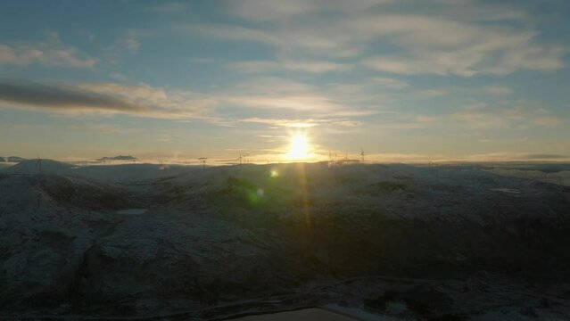 aerial view of a snu rising over mountain covered with snow with wind turbines on top of it in norway