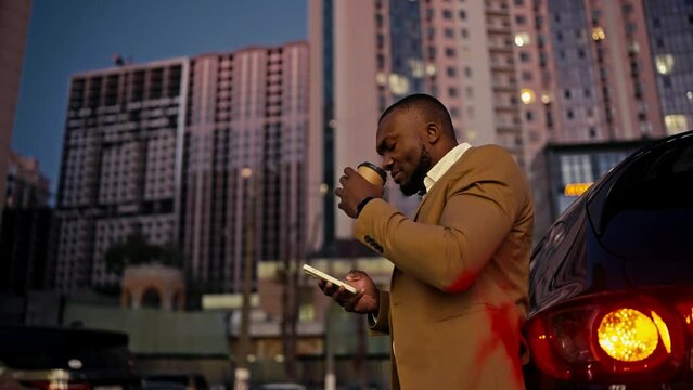 A confident and happy man with Black skin a short haircut and a beard in a brown suit stands near his black modern car drinks coffee and uses a white telephone in a city with large buildings in the