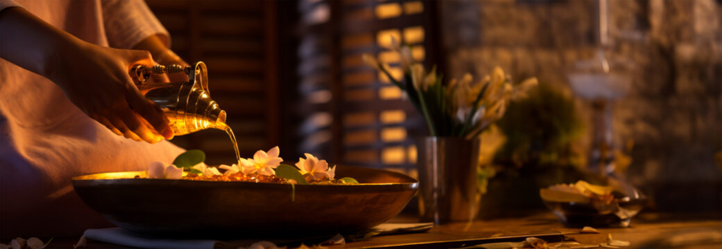In the soft illumination of evening, hands gracefully pour water from an ornate jug into a bowl with floating blossoms, a ritual purification and serenity in a tranquil spa setting. Ultra wide banner