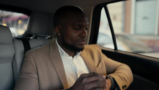 Confident man Businessman with Black skin and a beard in a brown suit rides in a modern car looks at his watch and then out the window. Successful businessman driving in a car to an appointed place
