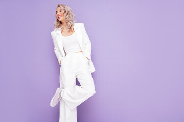 Portrait of young beautiful blond woman wearing nice trendy white suit. Fashion model posing in studio. Fashionable female isolated on violet background. Cheerful and happy