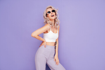 Naklejka premium Young beautiful smiling female in trendy summer white top and pants clothes. Carefree blond woman posing near violet wall in studio. Positive model having fun. Cheerful and happy. In sunglasses