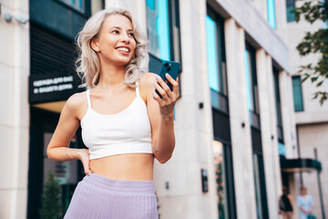 Young beautiful smiling hipster woman in trendy summer clothes. Carefree model posing in street at sunset. Positive blond female. Holds smartphone, uses phone apps, looks at telephone screen