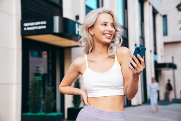 Young beautiful smiling hipster woman in trendy summer clothes. Carefree model posing in street at sunset. Positive blond female. Holds smartphone, uses phone apps, looks at telephone screen