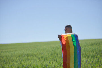Non-binary person, young and South American, very makeup, with a gay pride flag on his back, in the middle of a field of green wheat. Queen of the concept, lgbtq+, pride, queer.