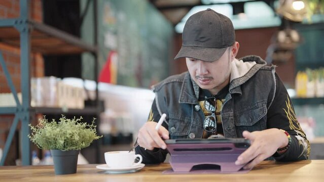 A Asian man working in a contemporary coffee shop drawing a picture and using a pen and digital tablet.