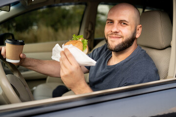 A bald smiling man driving a car drinking takeaway coffee and eating a burger looks out the window. Dangerous driving - obrazy, fototapety, plakaty