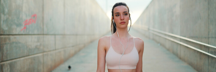 Young athletic woman with long ponytail wearing beige sports top with wired headphones walks along...